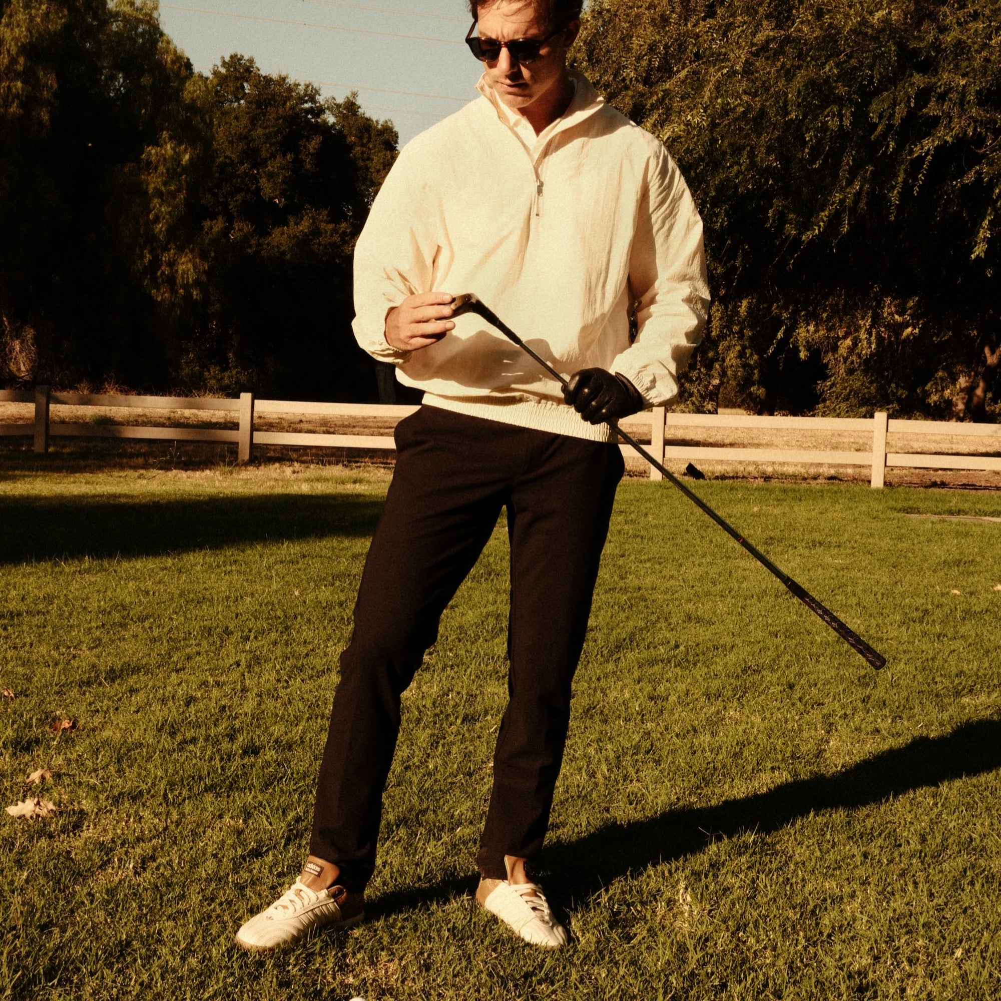 Luxury Western Golf apparel Man in a white shirt and sunglasses holding a golf club on a grassy field with trees in the background.