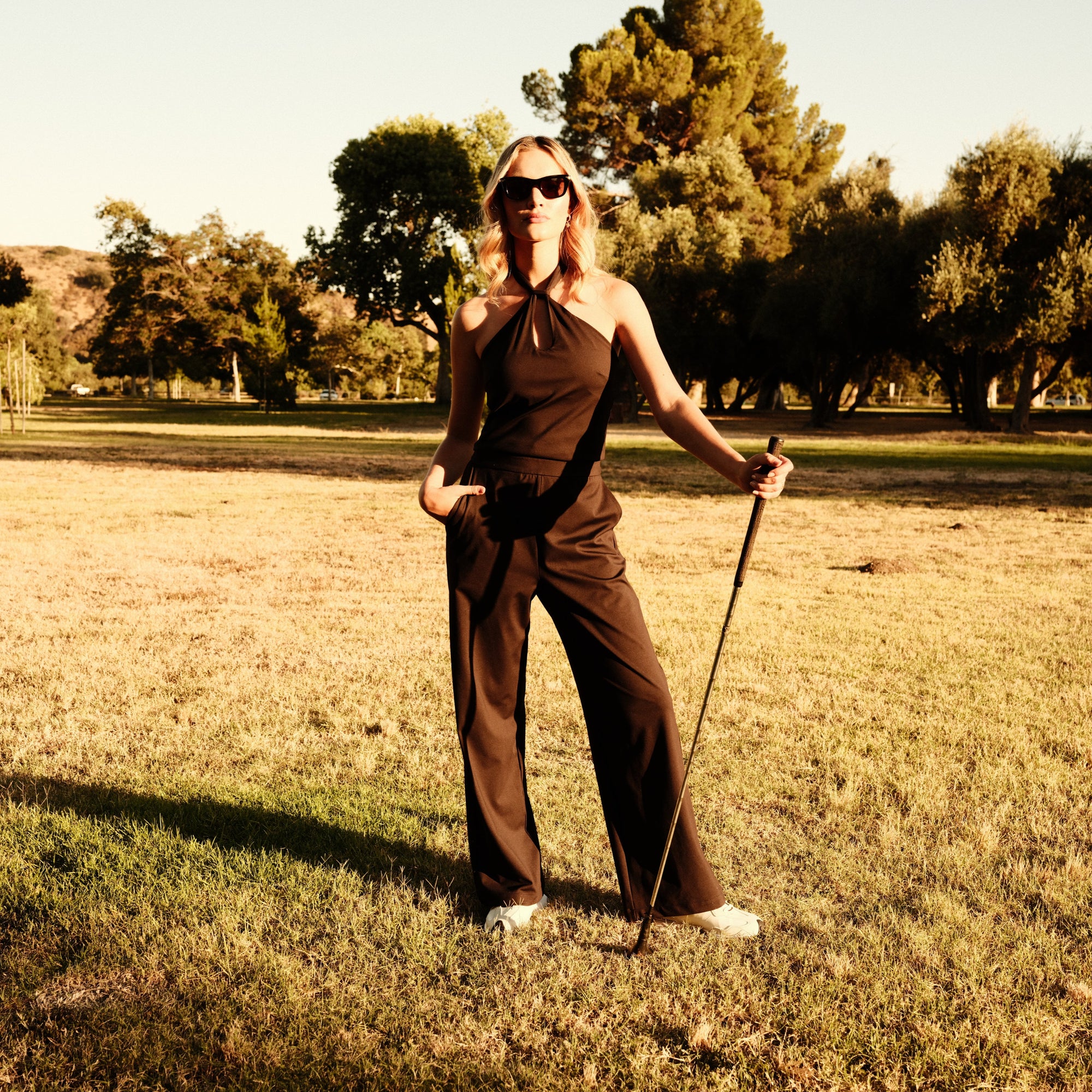 Luxury Western Golf apparel Woman in a black two piece jumpsuit standing in a grassy field with trees in the background