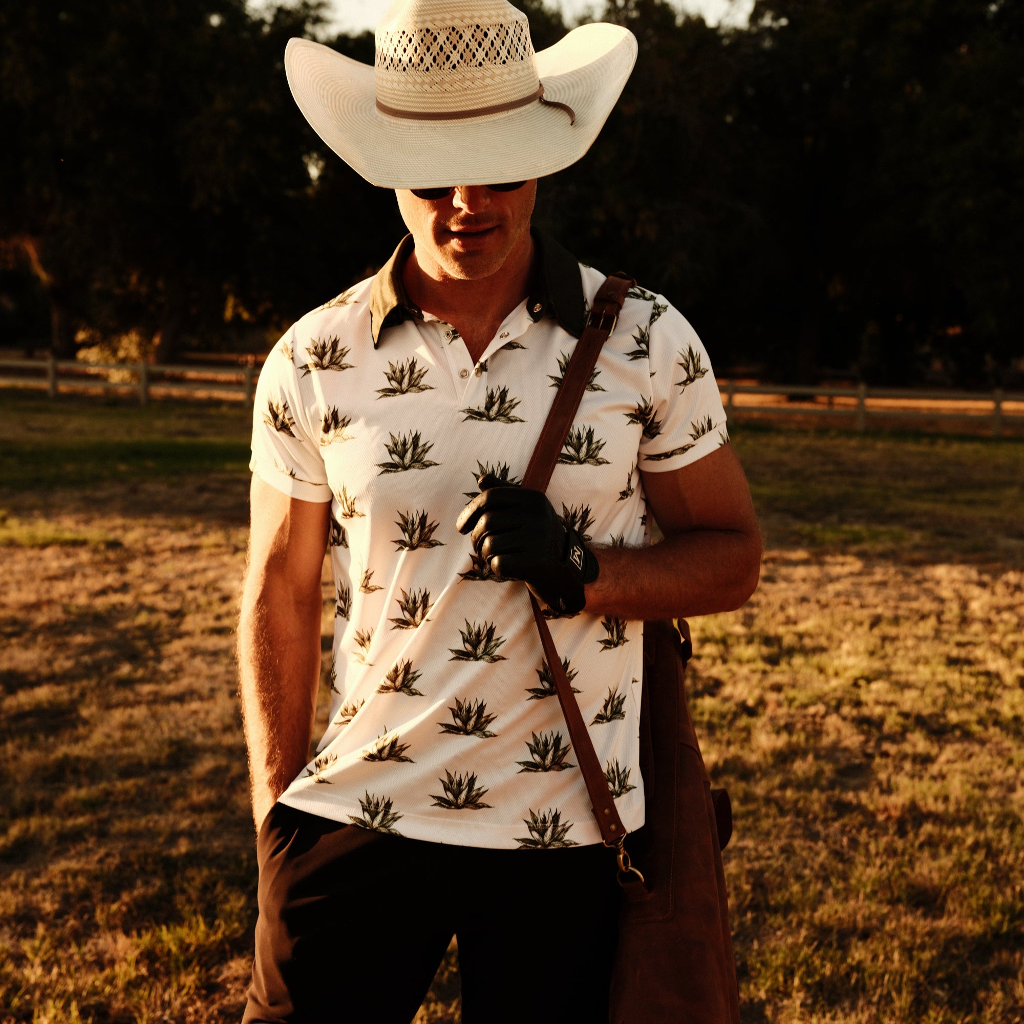 Luxury Western Golf apparel man wearing golf polo with agave print and a cowboy hat while holding a leather golf bag.