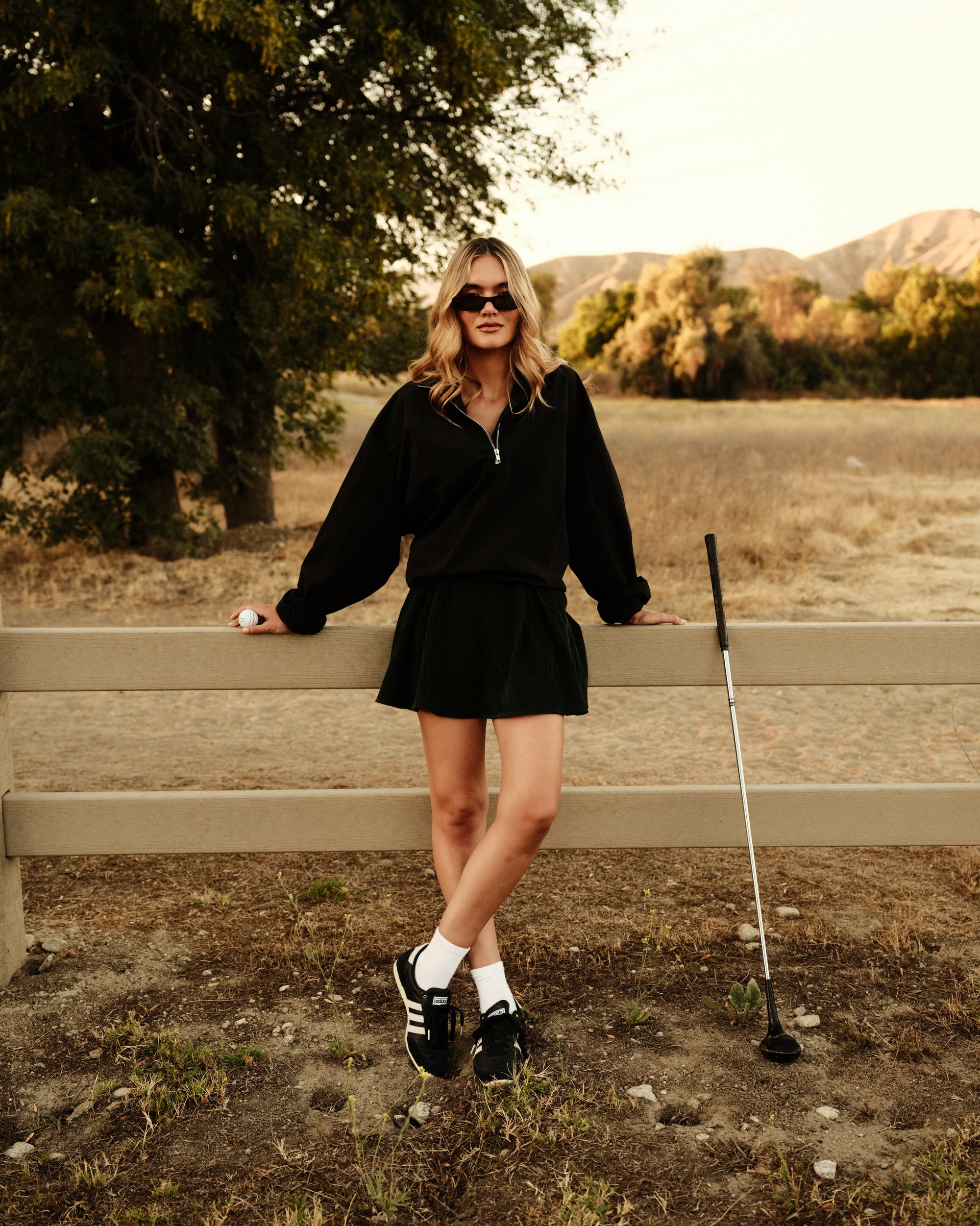 Woman in black outfit standing by a wooden fence with trees and mountains in the background