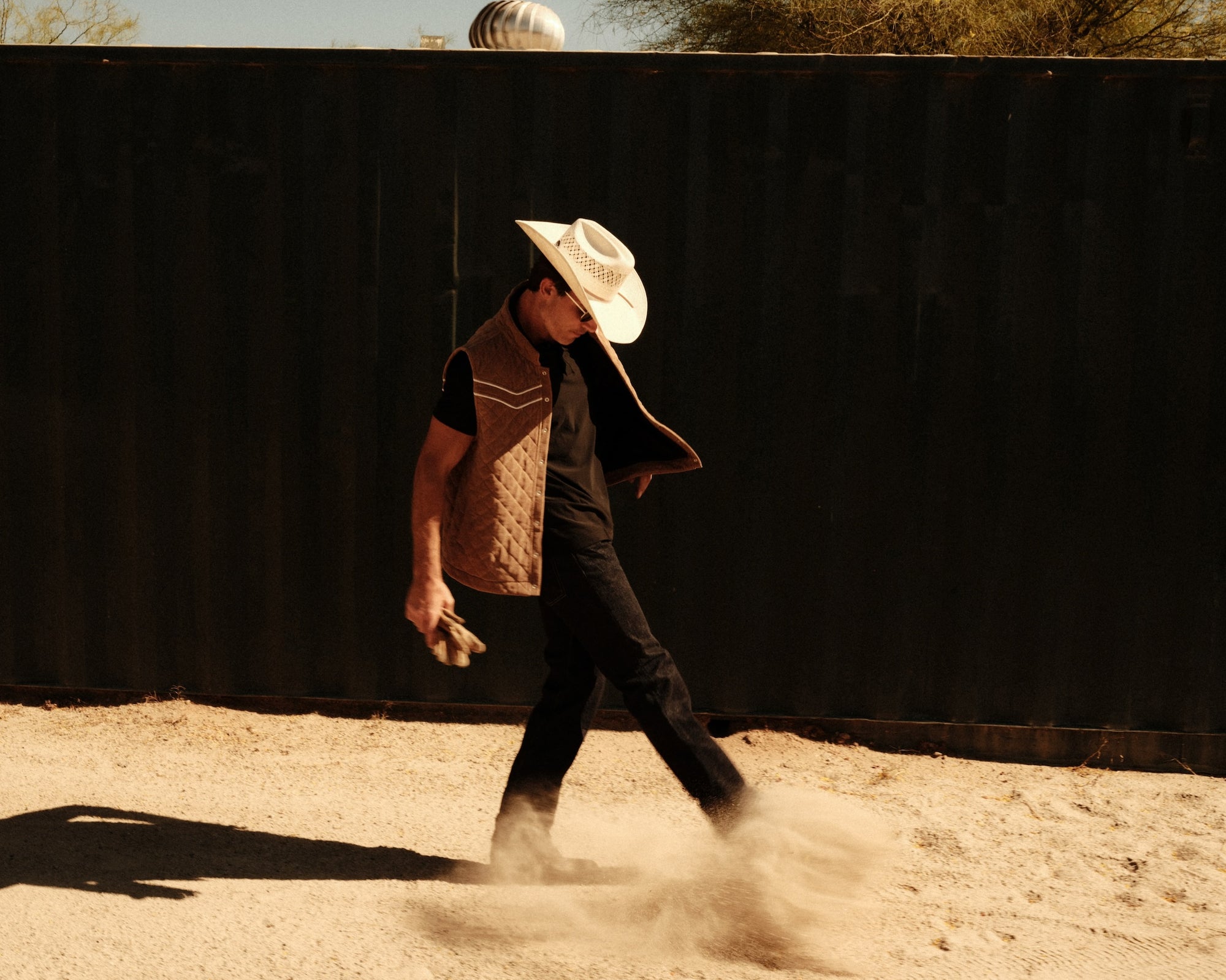 Person in cowboy hat and vest walking on a dirt path with a black wall in the background