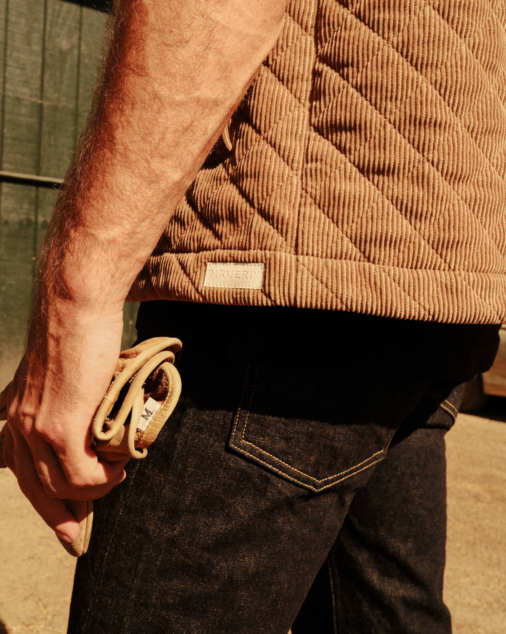 Person wearing a brown quilted vest and dark jeans, holding a leather belt.