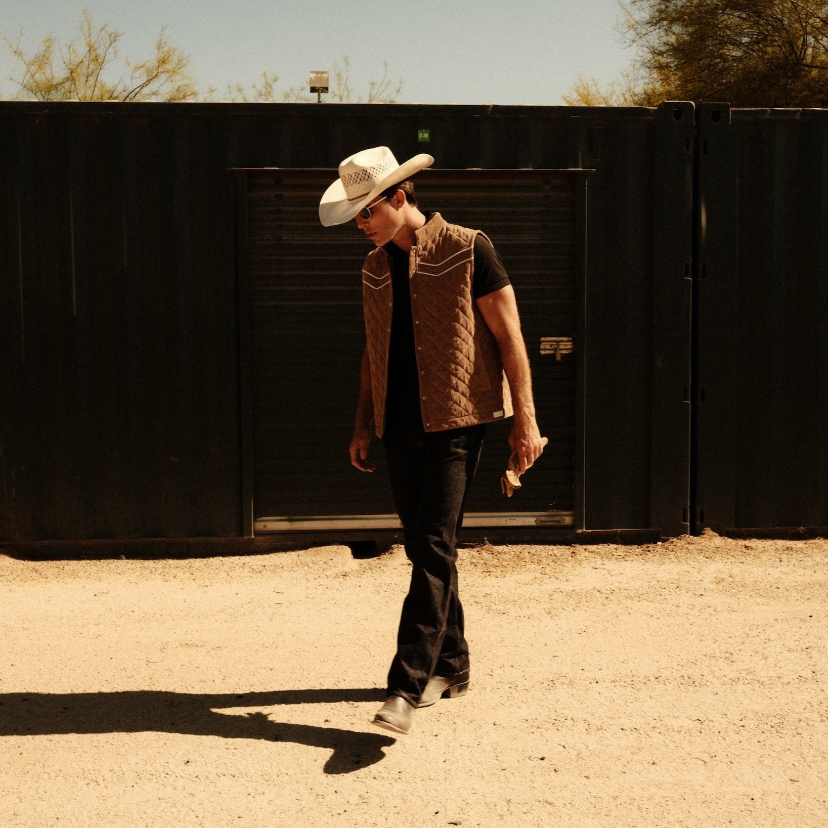Luxury Western Golf apparel Person wearing a cowboy hat and vest walking on a dirt path with a black fence and trees in the background.